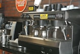 A commercial espresso machine is placed on a counter, equipped with multiple group heads and steam wands. Beside it are stacks of black disposable cups. The brand 'Café Bustelo' is prominently featured on the machine. A metal milk frothing pitcher and other coffee-making equipment are visible, set against a backdrop of a red brick wall with a Hatuey sign.