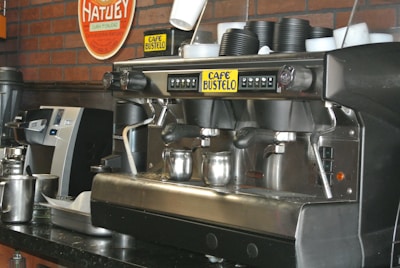 A commercial espresso machine is placed on a counter, equipped with multiple group heads and steam wands. Beside it are stacks of black disposable cups. The brand 'Café Bustelo' is prominently featured on the machine. A metal milk frothing pitcher and other coffee-making equipment are visible, set against a backdrop of a red brick wall with a Hatuey sign.