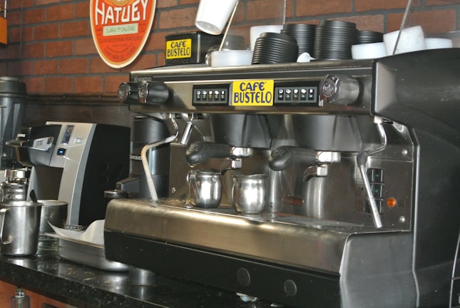 A commercial espresso machine is placed on a counter, equipped with multiple group heads and steam wands. Beside it are stacks of black disposable cups. The brand 'Café Bustelo' is prominently featured on the machine. A metal milk frothing pitcher and other coffee-making equipment are visible, set against a backdrop of a red brick wall with a Hatuey sign.