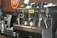 A commercial espresso machine is placed on a counter, equipped with multiple group heads and steam wands. Beside it are stacks of black disposable cups. The brand 'Café Bustelo' is prominently featured on the machine. A metal milk frothing pitcher and other coffee-making equipment are visible, set against a backdrop of a red brick wall with a Hatuey sign.