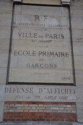 A stone wall with inscriptions in French, including 'Ville de Paris, École Primaire de Garçons, 1908' and 'Défense d'Afficher.' It also features the phrase 'Liberté, Égalité, Fraternité' and a date reference of 29 Juillet 1881. Red and beige bricks are visible at the bottom.