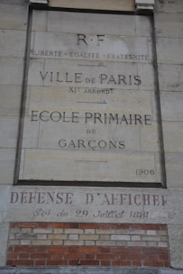 A stone wall with inscriptions in French, including 'Ville de Paris, École Primaire de Garçons, 1908' and 'Défense d'Afficher.' It also features the phrase 'Liberté, Égalité, Fraternité' and a date reference of 29 Juillet 1881. Red and beige bricks are visible at the bottom.