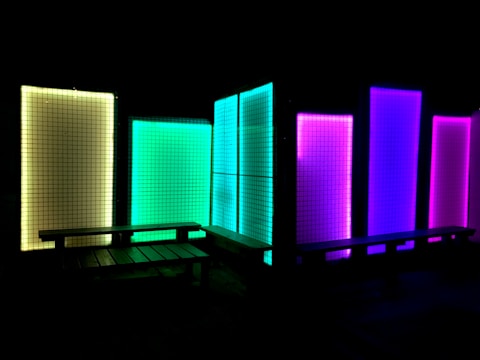 A series of illuminated rectangular panels display vibrant colors against a dark background. The panels are arranged in a sequence and emit hues of yellow, green, blue, and purple, creating a visually striking contrast with the surrounding darkness. In front of the panels are a couple of wooden benches, adding an element of seating to the scene.