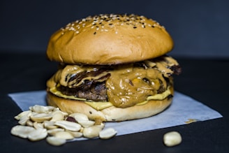 A juicy burger topped with a generous layer of creamy peanut butter sauce sits on a white paper sheet. The bun is sprinkled with sesame seeds, and a small pile of shelled peanuts is placed beside the burger. The background is a dark matte color, highlighting the textures and colors of the burger.