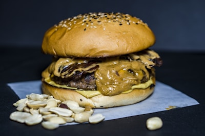 A juicy burger topped with a generous layer of creamy peanut butter sauce sits on a white paper sheet. The bun is sprinkled with sesame seeds, and a small pile of shelled peanuts is placed beside the burger. The background is a dark matte color, highlighting the textures and colors of the burger.
