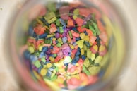 A collection of small, colorful pebbles or stones viewed through a transparent container. The stones are vibrant, featuring a mix of colors including pink, blue, green, yellow, and orange, creating a visually striking pattern.