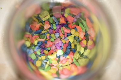 A collection of small, colorful pebbles or stones viewed through a transparent container. The stones are vibrant, featuring a mix of colors including pink, blue, green, yellow, and orange, creating a visually striking pattern.