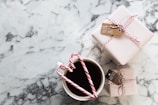 A marble table surface with two neatly wrapped gifts tied with red and white string and small tags. In the foreground, a cup filled with a dark beverage, adorned with two candy canes forming a heart shape.