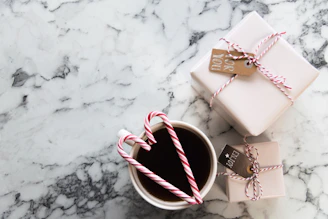 A marble table surface with two neatly wrapped gifts tied with red and white string and small tags. In the foreground, a cup filled with a dark beverage, adorned with two candy canes forming a heart shape.