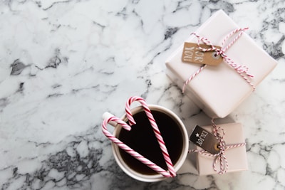 A marble table surface with two neatly wrapped gifts tied with red and white string and small tags. In the foreground, a cup filled with a dark beverage, adorned with two candy canes forming a heart shape.