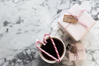 A marble table surface with two neatly wrapped gifts tied with red and white string and small tags. In the foreground, a cup filled with a dark beverage, adorned with two candy canes forming a heart shape.