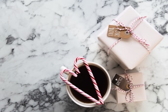 A marble table surface with two neatly wrapped gifts tied with red and white string and small tags. In the foreground, a cup filled with a dark beverage, adorned with two candy canes forming a heart shape.