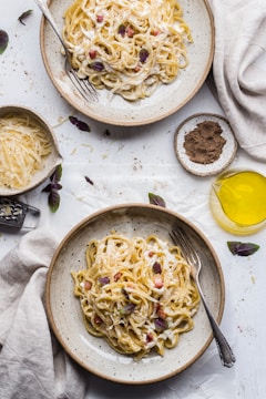 Two bowls of creamy pasta are placed on a table with scattered herbs and seasoning. The pasta is garnished with small bits of purple basil and pieces of bacon. Nearby, there are bowls containing grated cheese and ground pepper. A fork is resting in each bowl of pasta. The setup also includes a glass jar of olive oil and a light, rustic tablecloth.