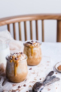 Two glass jars filled with creamy dessert topped with caramel sauce, oats, and chocolate chips are placed on a white surface. Surrounding them are scattered oats and chocolate pieces, some vintage spoons, and a small plate with cinnamon. The background features a wooden chair and soft fabric.