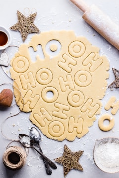 A flat surface is covered with rolled-out cookie dough featuring cutout letters spelling 'HO HO HO'. Surrounding the dough are baking items, including a rolling pin, a bowl of flour, measuring spoons, star-shaped ornaments, and kitchen twine. The composition conveys a festive baking theme.