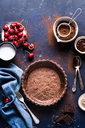A rustic baking setup featuring a prebaked chocolate crust placed centrally on a dark surface. Surrounding the crust are fresh cherries, some still with stems, in a small dish. A cup of milk and scattered cocoa powder add to the baking theme. Nearby are a sieve with cocoa and a bowl containing liquid chocolate.