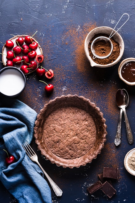 A rustic baking setup featuring a prebaked chocolate crust placed centrally on a dark surface. Surrounding the crust are fresh cherries, some still with stems, in a small dish. A cup of milk and scattered cocoa powder add to the baking theme. Nearby are a sieve with cocoa and a bowl containing liquid chocolate.