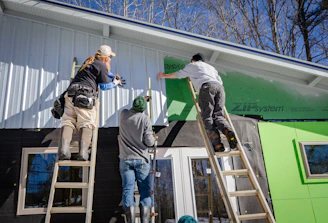 Three individuals are working on the exterior of a building, possibly engaged in construction or renovation. Two are standing on ladders, attaching or securing siding material to the upper part of the structure. The setting is outdoors in a sunny environment with leafless trees visible in the background.