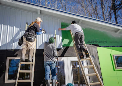 Three individuals are working on the exterior of a building, possibly engaged in construction or renovation. Two are standing on ladders, attaching or securing siding material to the upper part of the structure. The setting is outdoors in a sunny environment with leafless trees visible in the background.