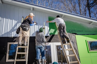 Three individuals are working on the exterior of a building, possibly engaged in construction or renovation. Two are standing on ladders, attaching or securing siding material to the upper part of the structure. The setting is outdoors in a sunny environment with leafless trees visible in the background.