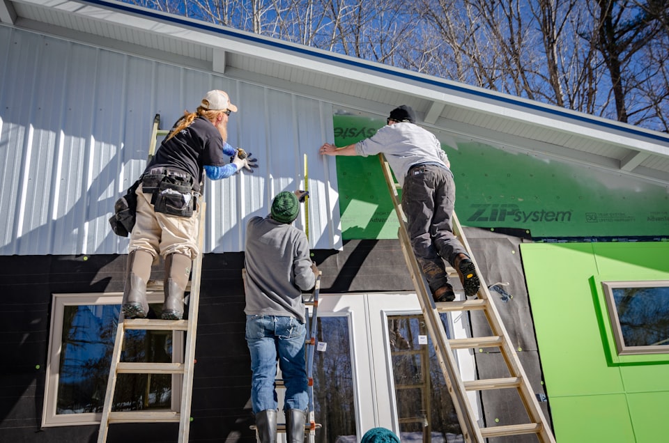 Three contractors on ladders installing siding over green sheathing on a New England home in winter, the kind of remodeling work that needs a Rhode Island contractor license