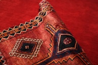 A richly patterned textile with a prominent geometric design featuring diamonds and intricate borders, primarily in deep red, black, and orange tones. The fabric is partially rolled, showcasing its texture and weaving patterns.