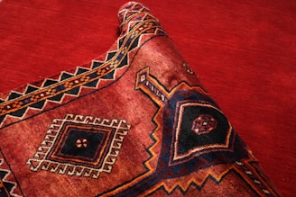 A richly patterned textile with a prominent geometric design featuring diamonds and intricate borders, primarily in deep red, black, and orange tones. The fabric is partially rolled, showcasing its texture and weaving patterns.