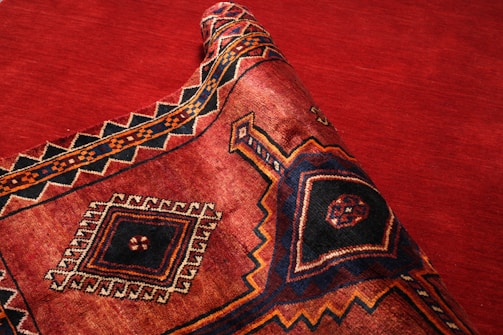 A richly patterned textile with a prominent geometric design featuring diamonds and intricate borders, primarily in deep red, black, and orange tones. The fabric is partially rolled, showcasing its texture and weaving patterns.