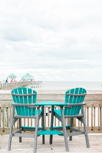 Two teal wooden chairs and a small matching table are placed on a wooden deck, overlooking a beach with a pier extending into the ocean. The sky is mostly cloudy, creating a serene and peaceful atmosphere.