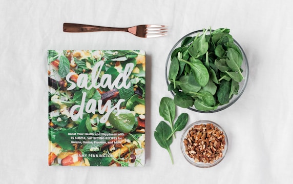 A cookbook titled 'Salad Days' is displayed on a white surface. Next to the book are a fork, a bowl of fresh spinach leaves, some loose spinach, and a small bowl of nuts.