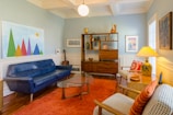 A cozy living room with mid-century modern design. It features a blue sofa, an orange shag rug, and a glass coffee table. There is a wooden bookshelf displaying various decorative items, including vases and art pieces. A guitar is leaned against the wall. The walls are painted light blue and decorated with vibrant artworks, including a colorful geometric painting.