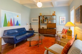 A cozy living room with mid-century modern design. It features a blue sofa, an orange shag rug, and a glass coffee table. There is a wooden bookshelf displaying various decorative items, including vases and art pieces. A guitar is leaned against the wall. The walls are painted light blue and decorated with vibrant artworks, including a colorful geometric painting.