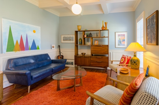 A cozy living room with mid-century modern design. It features a blue sofa, an orange shag rug, and a glass coffee table. There is a wooden bookshelf displaying various decorative items, including vases and art pieces. A guitar is leaned against the wall. The walls are painted light blue and decorated with vibrant artworks, including a colorful geometric painting.