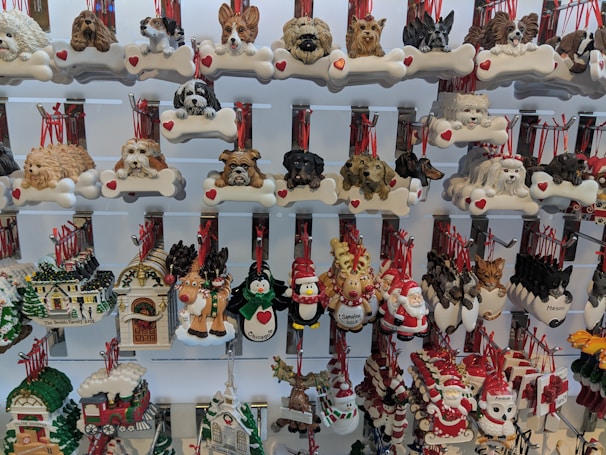 A display of various decorative ornaments, featuring a variety of dog breeds, and festive holiday symbols such as Santa Claus, reindeer, and snowmen. Each ornament includes detailed and colorful designs, many with red ribbons for hanging.