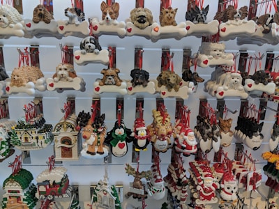 A display of various decorative ornaments, featuring a variety of dog breeds, and festive holiday symbols such as Santa Claus, reindeer, and snowmen. Each ornament includes detailed and colorful designs, many with red ribbons for hanging.