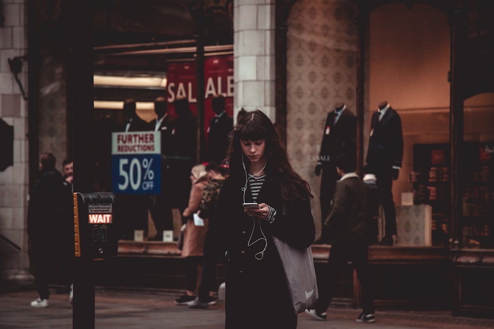 donna in piedi che indossa cappotto nero usando smartphone e auricolari