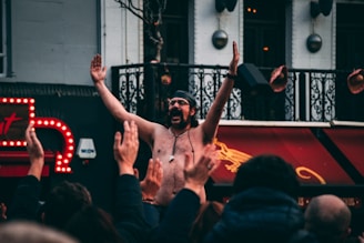 A shirtless man wearing a headband and sunglasses raises his arms in celebration or excitement amidst a crowd of people with raised hands. The setting appears to be urban, with a backdrop featuring ornate railings and bright neon signage. The crowd seems engaged and lively.