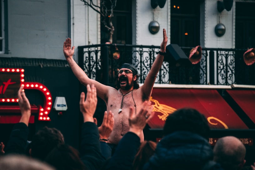A shirtless man wearing a headband and sunglasses raises his arms in celebration or excitement amidst a crowd of people with raised hands. The setting appears to be urban, with a backdrop featuring ornate railings and bright neon signage. The crowd seems engaged and lively.