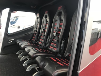 The image depicts the interior of a vehicle featuring a row of four black leather seats with red accents and harness-style seat belts. The seats are aligned side by side on a dark carpeted floor, and there is a window providing an outside view.