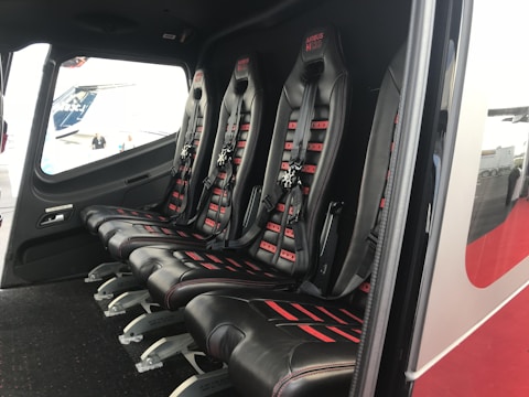 The image depicts the interior of a vehicle featuring a row of four black leather seats with red accents and harness-style seat belts. The seats are aligned side by side on a dark carpeted floor, and there is a window providing an outside view.