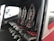 The image depicts the interior of a vehicle featuring a row of four black leather seats with red accents and harness-style seat belts. The seats are aligned side by side on a dark carpeted floor, and there is a window providing an outside view.