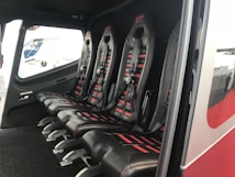 The image depicts the interior of a vehicle featuring a row of four black leather seats with red accents and harness-style seat belts. The seats are aligned side by side on a dark carpeted floor, and there is a window providing an outside view.