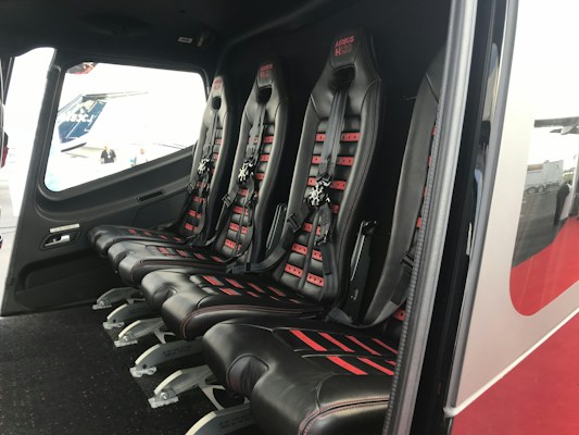 The image depicts the interior of a vehicle featuring a row of four black leather seats with red accents and harness-style seat belts. The seats are aligned side by side on a dark carpeted floor, and there is a window providing an outside view.