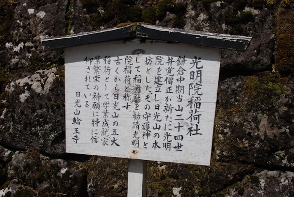 A wooden sign features vertical Japanese characters, set against a textured stone wall with visible moss patches.