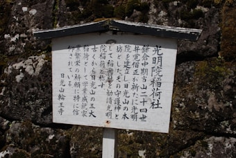 A wooden sign features vertical Japanese characters, set against a textured stone wall with visible moss patches.