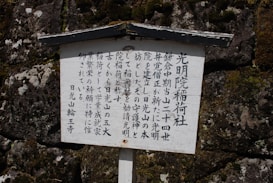 A wooden sign features vertical Japanese characters, set against a textured stone wall with visible moss patches.