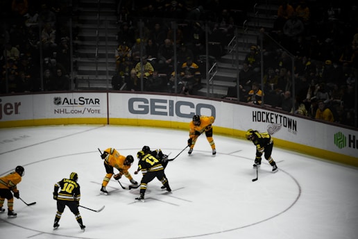 A group of ice hockey players in yellow and black jerseys are engaged in a face-off on the rink. The ice is white, and the arena is filled with spectators dressed in similar colors, indicating team support. Various advertisements are visible on the perimeter boards.