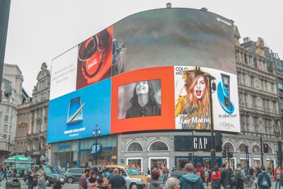 A busy city square featuring a massive digital billboard displaying various advertisements, including brands like Coca-Cola, Samsung, and L'Oreal. The advertisements show images of products and people, creating a bright and lively atmosphere. The surrounding area is bustling with people walking and vehicles moving through the street.