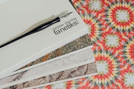 A stack of materials, possibly sample cards or tiles, displayed on top of a patterned background. The stack is topped with a plain card featuring the text 'Fanetik Marble Design Studio' and a black pen resting on it. The patterned background consists of intricate geometric designs in vibrant red, yellow, and green colors.