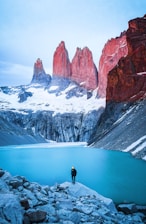 man standing on rock facing body of water and mountain